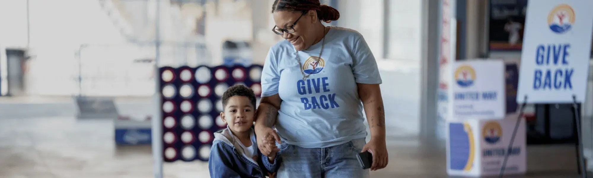 Young woman wearing blue Give Back shirt walking with small child