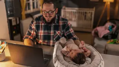 Father on laptop with infant resting in bed